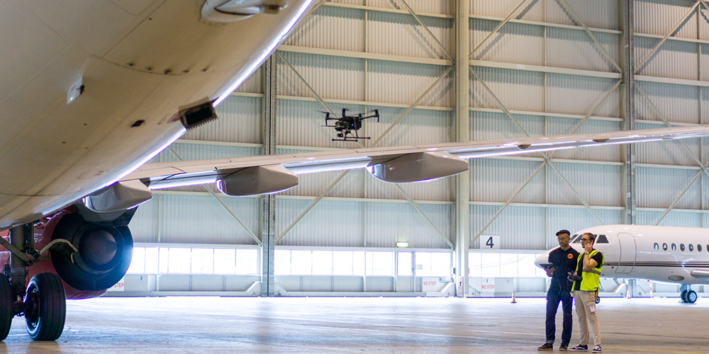 A hangar with aeroplanes and two people inspecting them with a drone