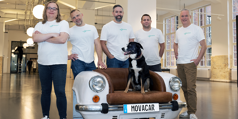 A team of people wearing white shirts, posing for a photo next to a dog sitting on a car-shaped sofa